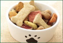 A white bowl with a black paw print contains assorted dog treats in various shapes, including bones and hearts.