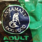 Close-up of a black and green "ANNAMAET DOG FOOD" bag labeled "ADULT" with 23% protein, featuring chicken meal and brown rice ingredients.