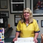 A woman wearing a yellow shirt stands behind a counter with a binder open, smiling. The background features framed pictures, certificates, and a wall-mounted touch screen device.