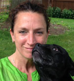 A person with dark hair pulled back, wearing a green shirt, smiles while a black dog nuzzles their face. They are outdoors with grass and a wooden fence in the background.