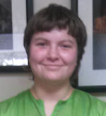 A person with short hair, wearing a green shirt, smiles in a close-up photo. Framed pictures are seen in the blurred background.